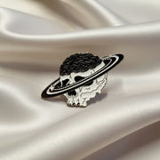 This enamel lapel pin features a human skull with its brain exposed, encircled by planetary rings on a light background