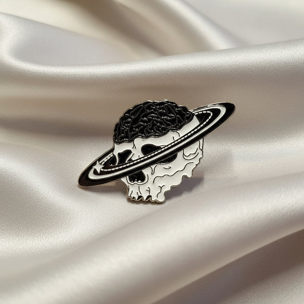 This enamel lapel pin features a human skull with its brain exposed, encircled by planetary rings on a light background