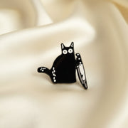 A pin depicting a wide-eyed black cat clutching a butcher knife on a light colored background.