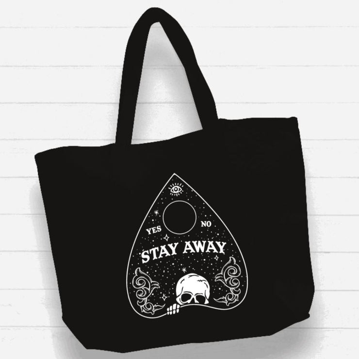Black tote bag with white planchette design and "Stay Away" text on white wood background