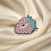 Strawberry-shaped pin with pink body and teal blue leaves on a beige background