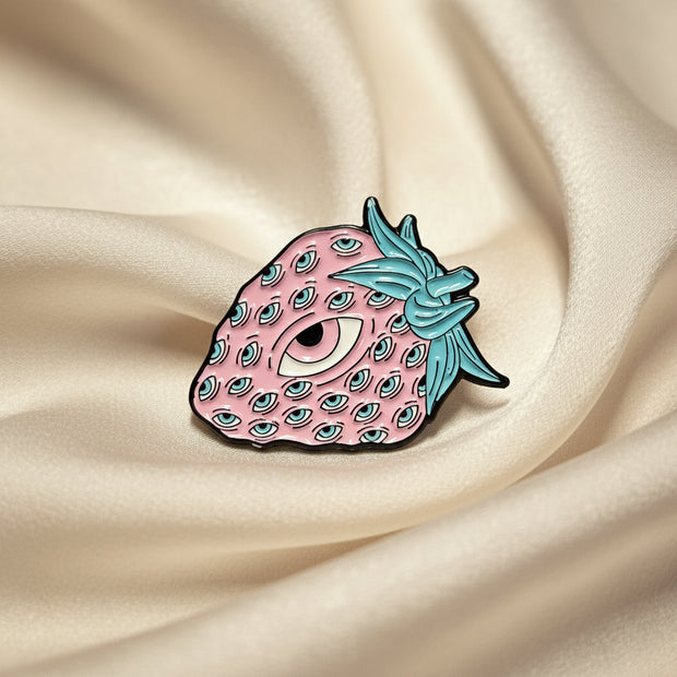 Strawberry-shaped pin with pink body and teal blue leaves on a beige background