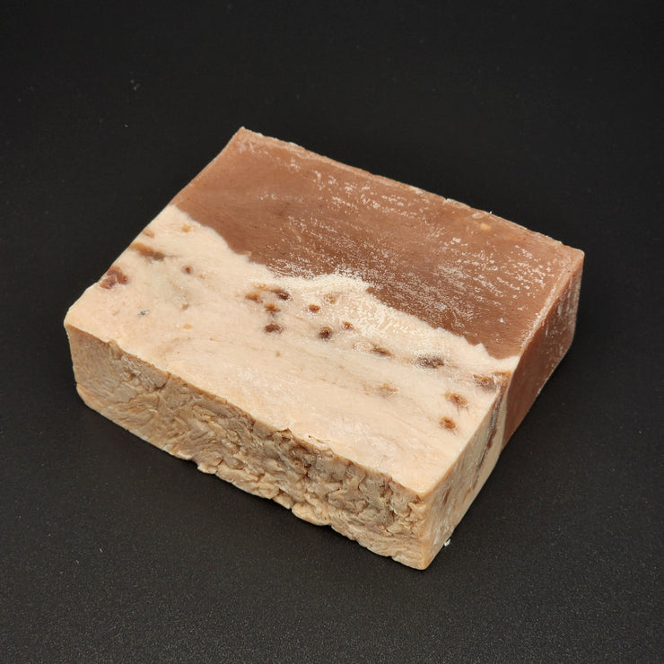 A rectangular bar of Sweet Tooth Handmade Vegan Bar Soap by Bleak Boutique, with a brown top and larger white base flecked with colorful dots, is shown on a smooth black surface. SEASONAL.