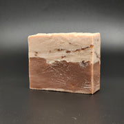 A rectangular bar of Sweet Tooth Handmade Vegan Bar Soap by Bleak Boutique, with a brown top and larger white base flecked with colorful dots, is shown on a smooth black surface. SEASONAL.