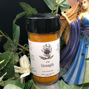 Tarot Card Spice Blend "Strength" by Dahlia's Enchanted Kitchen surrounded by greenery and next to a fairy statue.