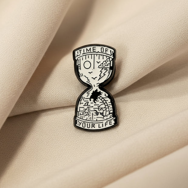 Black and white hourglass-shaped enamel pin with text on a light background