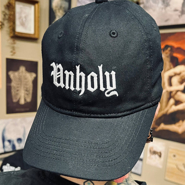 Black cap with 'Unholy' text worn by a person in a room with artistic decorations.