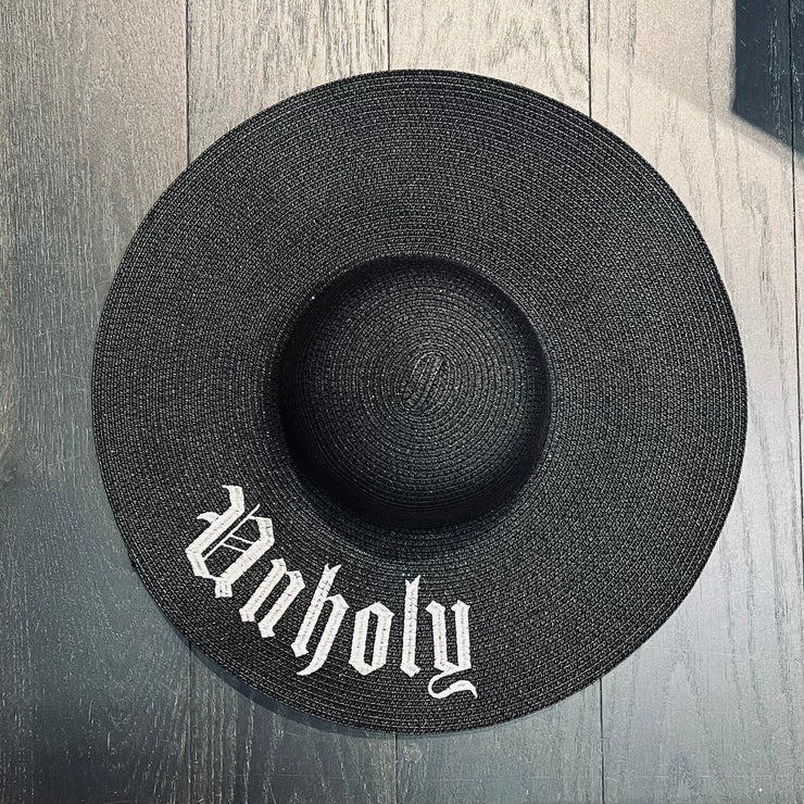 Black straw hat with 'Unholy' text on a wooden surface