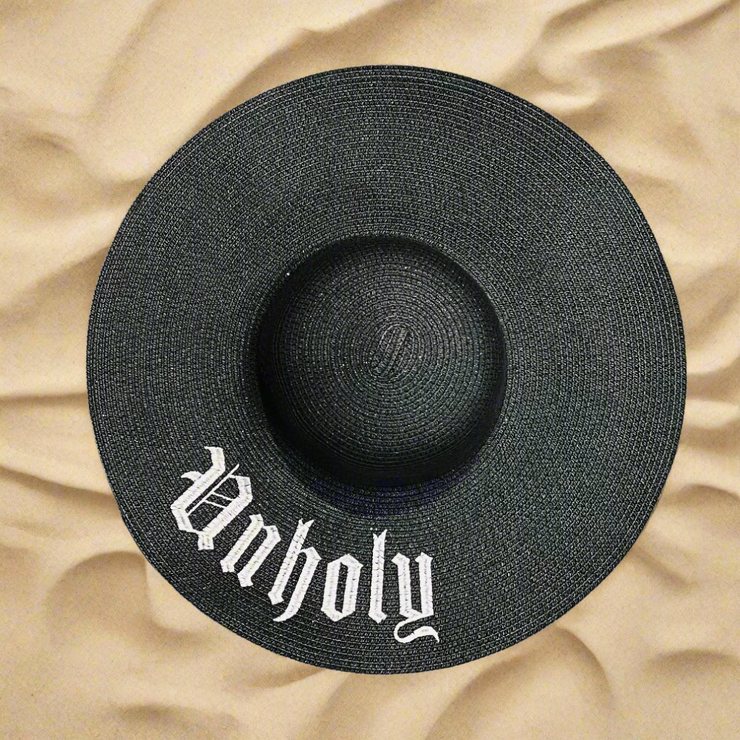 Black straw hat with 'Unholy' text on a beach sand background