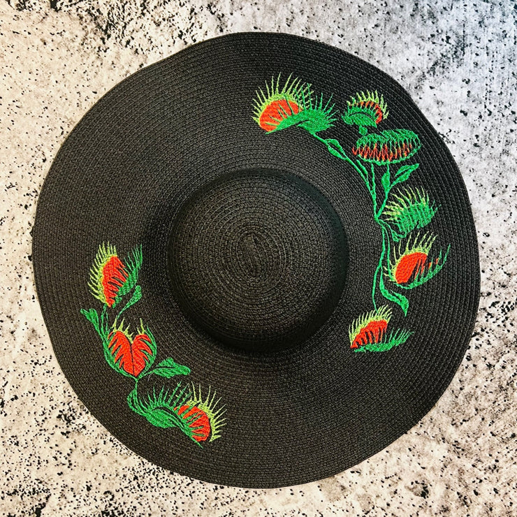 Black straw hat with red and green Venus fly trap embroidery on a textured surface