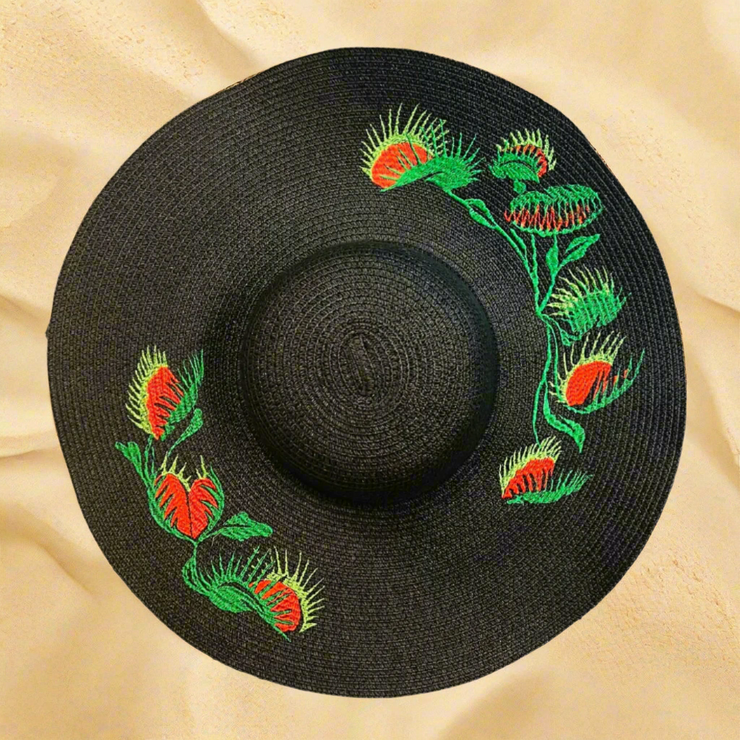 Black straw hat with green and red floral embroidery on a beach sand background
