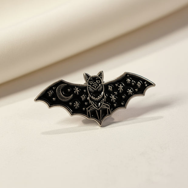 Bat shaped enamel pin with moon and star detailing on its wings on a light background