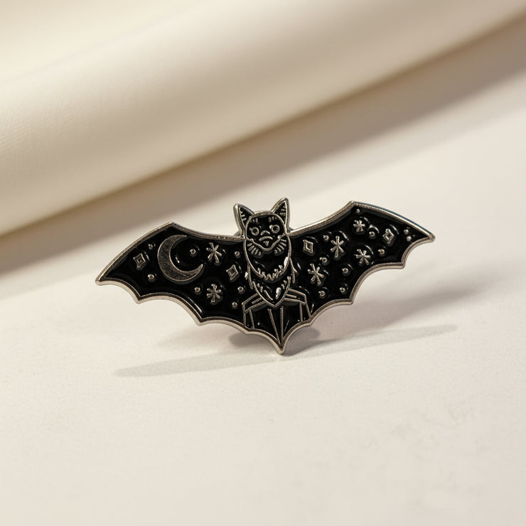 Bat shaped enamel pin with moon and star detailing on its wings on a light background