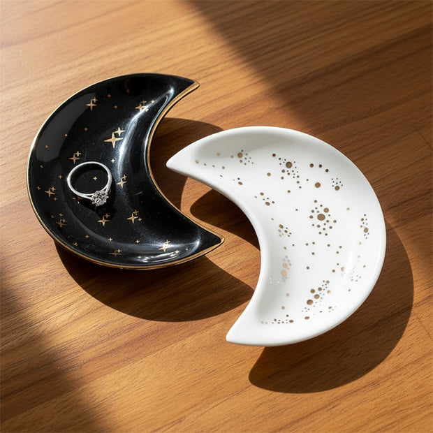 The Crescent Moon Ceramic Jewelry Valet Dish by Bleak Boutique features two interlocking pieces—one black with gold stars, one white with gold dots—forming a circle. The black dish holds jewelry, shown displaying a silver gemstone ring.