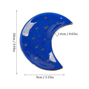 The Crescent Moon Ceramic Jewelry Valet Dish by Bleak Boutique is a blue, crescent-shaped ceramic dish with gold stars, ideal as a jewelry caddy or trinket holder. Size: 10 cm tall, 8 cm wide, 1.6 cm deep.