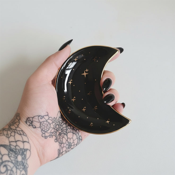 A hand displays the Bleak Boutique Crescent Moon Ceramic Jewelry Valet Dish, adorned with gold stars and edging—ideal as a trinket holder or jewelry caddy—set against a simple light background.
