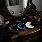 Three Crescent Moon Ceramic Jewelry Valet Dishes by Bleak Boutique—black, blue, and white with gold star patterns—are displayed on a dressing table. 